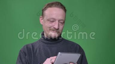 穿灰色套头衫的金发养老金领取者<strong>专心</strong>地工作在平板电脑上，并在绿色背景上观看相机。